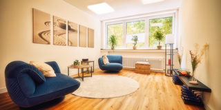 Ein Wohnzimmer mit Holzboden, zwei blauen Sesseln, einem runden Teppich, einem kleinen Tisch, Pflanzen auf der Fensterbank und Wandbildern.