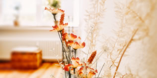 Ein Strau&szlig; getrockneter Blumen in warmen Farben steht in einem Raum mit Holzboden und einem unscharfen Fenster im Hintergrund.