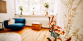 Ein Wohnzimmer mit einem blauen Sessel, einem Korbhocker, Holzboden und Pflanzen auf der Fensterbank. Im Vordergrund sind getrocknete Blumen zu sehen.