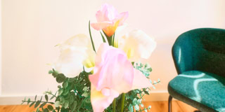 Ein Blumenstrau&szlig; mit rosa und wei&szlig;en Bl&uuml;ten steht neben einem gr&uuml;nen Sessel auf einem Holzboden vor einer wei&szlig;en Wand.