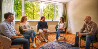 F&uuml;nf Personen sitzen in einem Kreis auf St&uuml;hlen in einem Raum mit einem gro&szlig;en Fenster im Hintergrund.