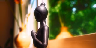 Schwarze Skulptur einer stehenden Figur vor einem Fenster mit unscharfem Hintergrund aus gr&uuml;nen B&auml;umen und orangefarbenen Vasen.