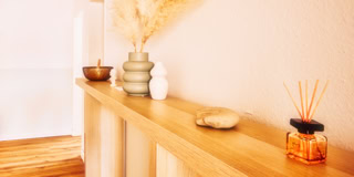 Holzregal mit Dekorationen, darunter eine Vase mit getrockneten Gr&auml;sern, eine kleine wei&szlig;e Skulptur, ein Stein und ein Glas mit Duftst&auml;bchen.