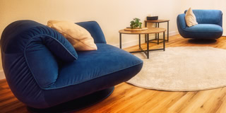Zwei blaue Sessel mit Kissen auf einem Holzboden, ein runder Teppich und ein kleiner Tisch mit Pflanzen und B&uuml;chern.