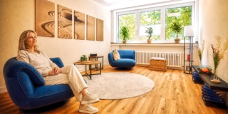 Eine Person sitzt auf einem blauen Sessel in einem hellen Raum mit Holzboden, einem runden Teppich, Pflanzen auf der Fensterbank und Wanddekorationen.