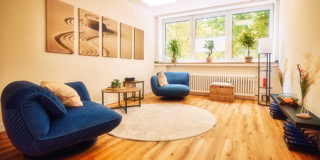 Ein Wohnzimmer mit Holzboden, zwei blauen Sesseln, einem runden Teppich, einem kleinen Tisch, Pflanzen auf der Fensterbank und Wandbildern.